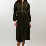 A woman modeling a dark olive green two-piece outfit consisting of a cropped zip-front jacket and a matching midi skirt, paired with beige pointed-toe heels against a light gray studio background.