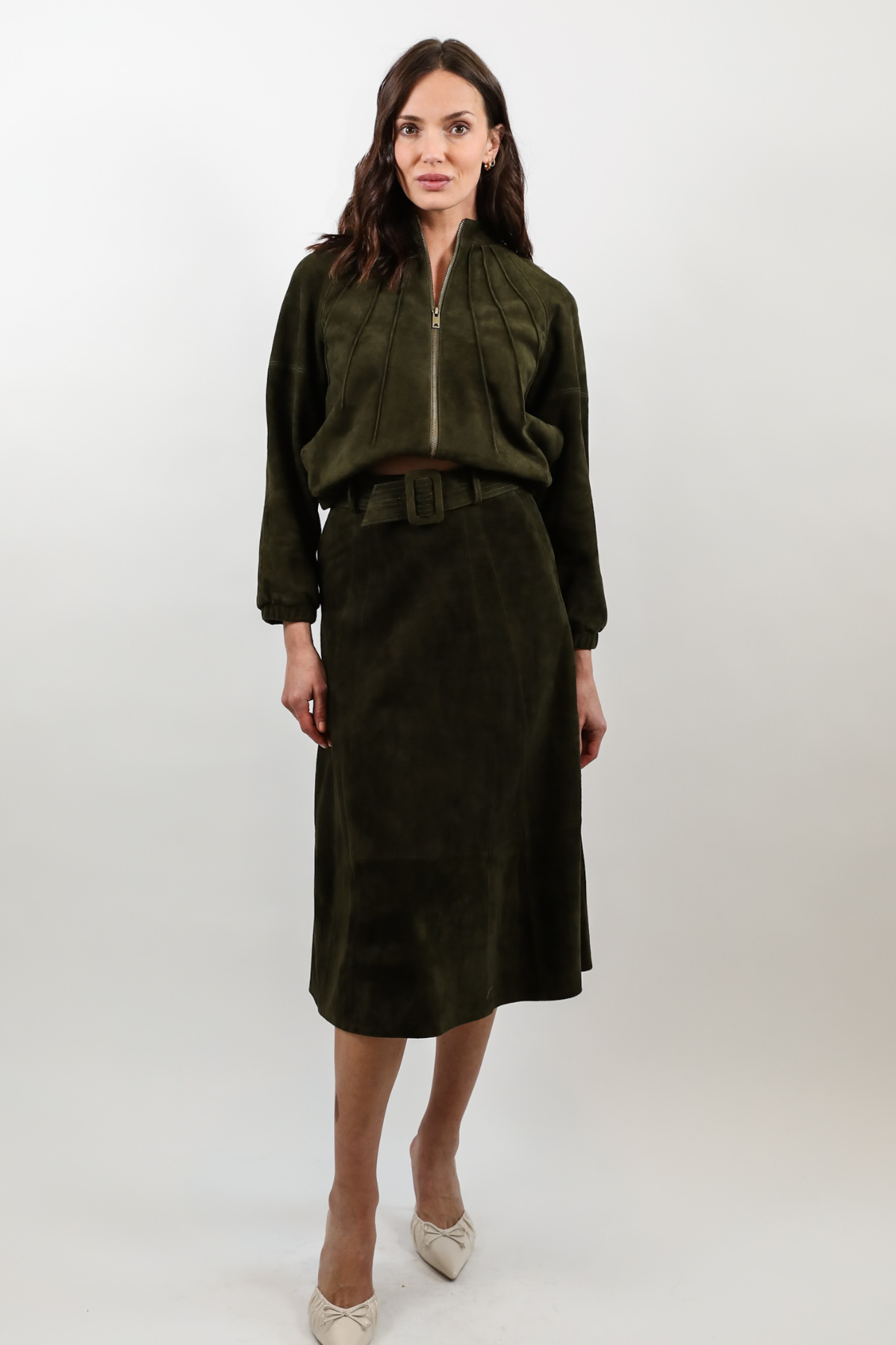 A woman modeling a dark olive green two-piece outfit consisting of a cropped zip-front jacket and a matching midi skirt, paired with beige pointed-toe heels against a light gray studio background.