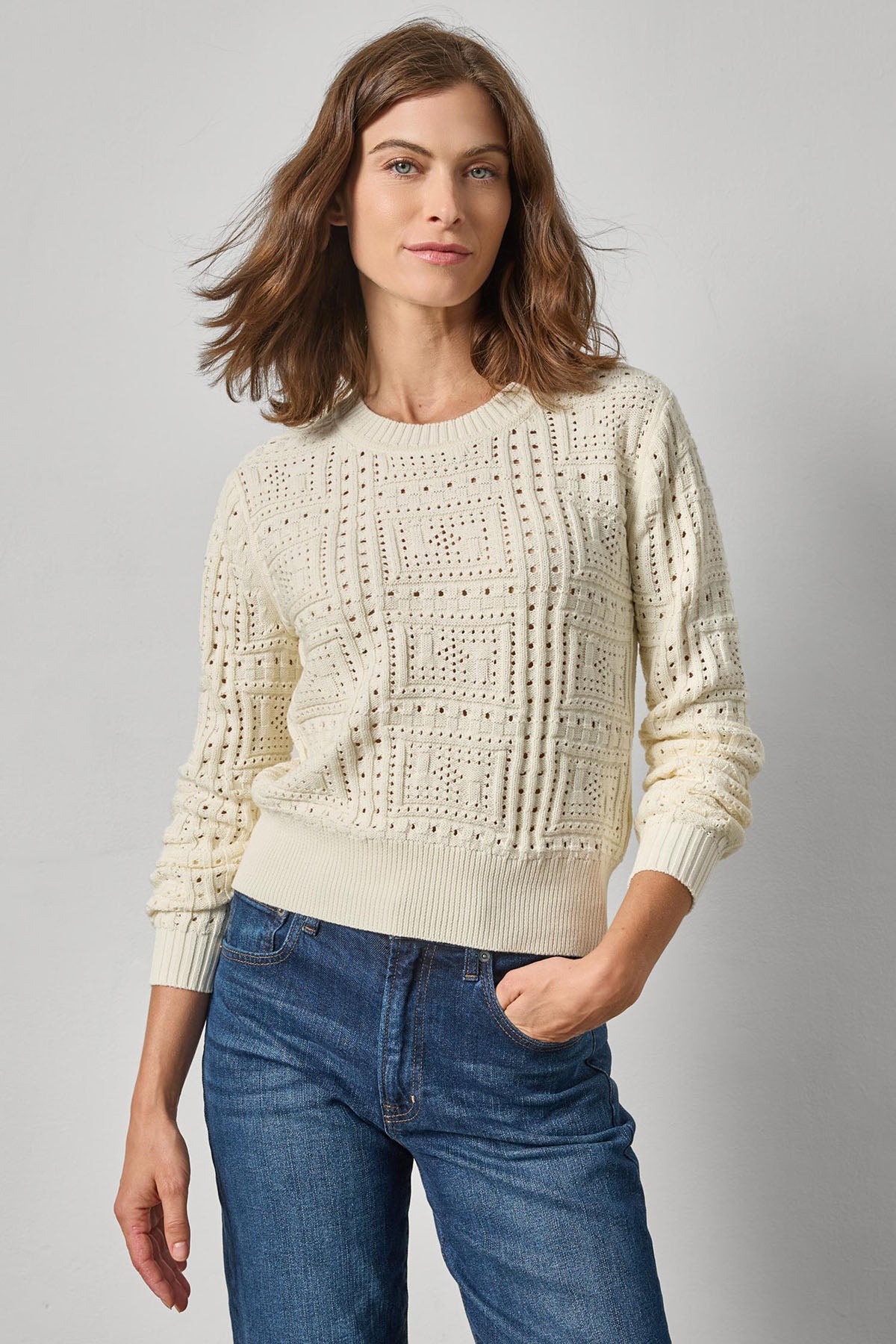 Woman wearing a cream knitted sweater and blue jeans against a gray background