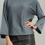 A woman wearing a slate blue long-sleeve knit top with wide bell sleeves and black faux-leather pants, shown from the lips down against a plain light gray studio background.