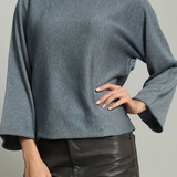 A woman wearing a slate blue long-sleeve knit top with wide bell sleeves and black faux-leather pants, shown from the lips down against a plain light gray studio background.