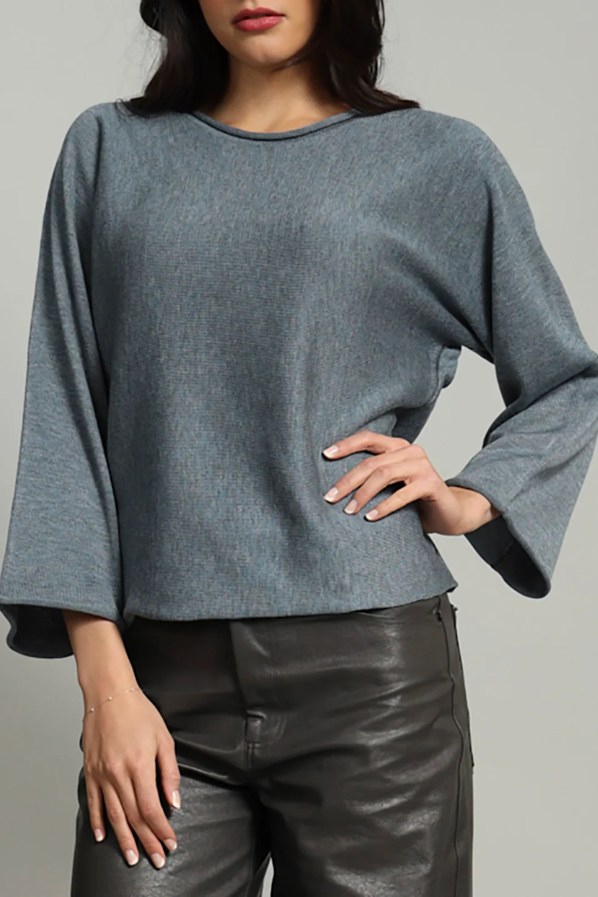 A woman wearing a slate blue long-sleeve knit top with wide bell sleeves and black faux-leather pants, shown from the lips down against a plain light gray studio background.