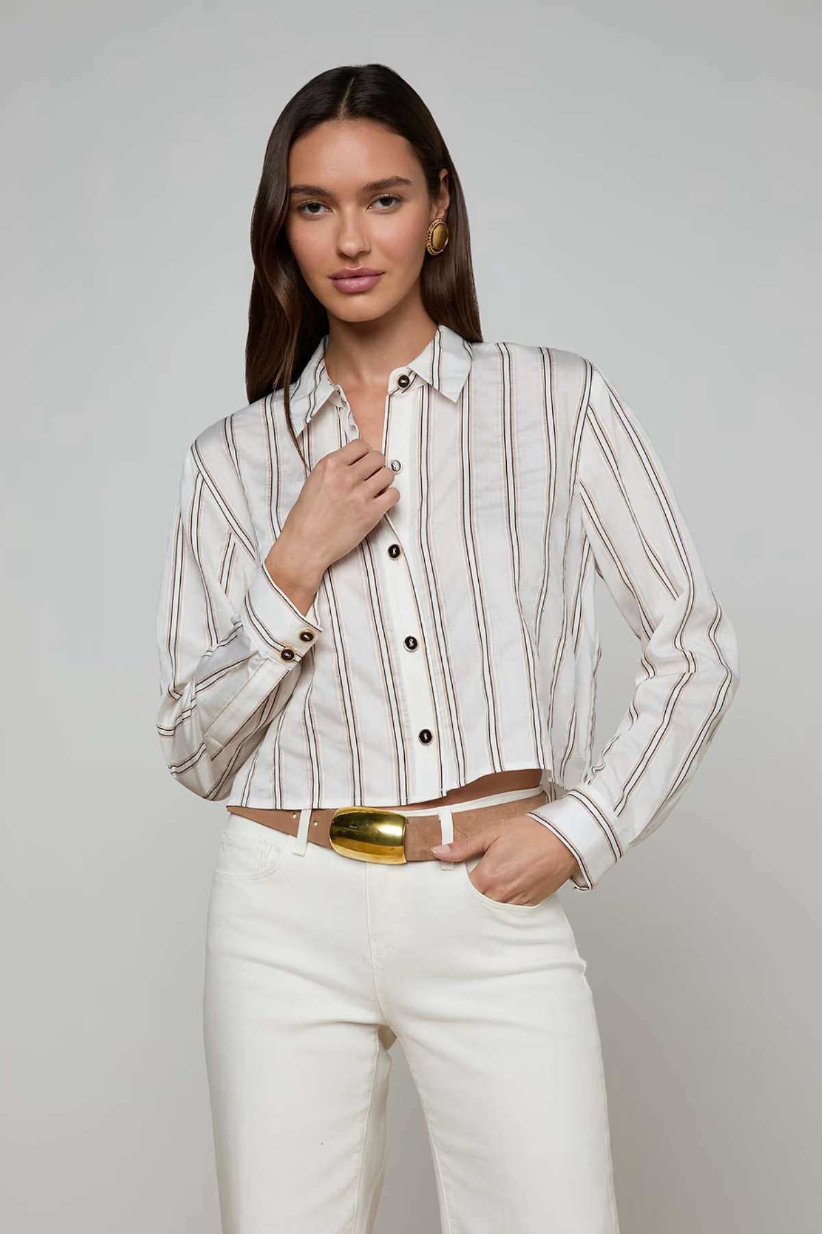 Woman wearing a striped shirt and white pants with a gold belt on a gray background