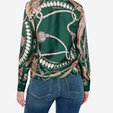 A woman shown from the back wearing a green satin shirt jacket with a detailed horse bit and rope print, paired with blue jeans, against a light gray studio background.