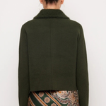 A model shown from the back wearing a dark olive green cropped jacket with a fuzzy collar and a patterned green, brown, and gold skirt against a plain white background.