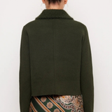 A model shown from the back wearing a dark olive green cropped jacket with a fuzzy collar and a patterned green, brown, and gold skirt against a plain white background.