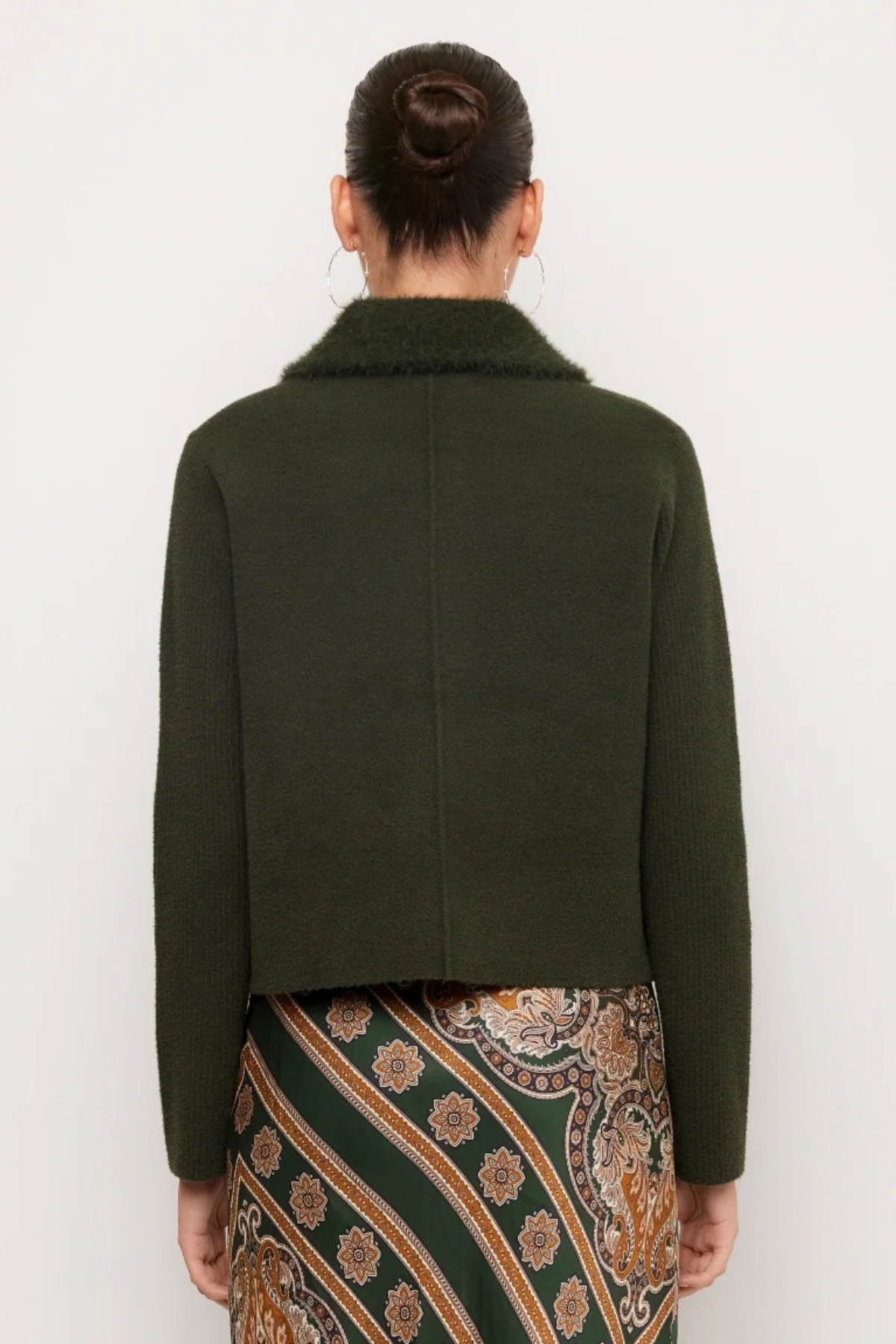 A model shown from the back wearing a dark olive green cropped jacket with a fuzzy collar and a patterned green, brown, and gold skirt against a plain white background.