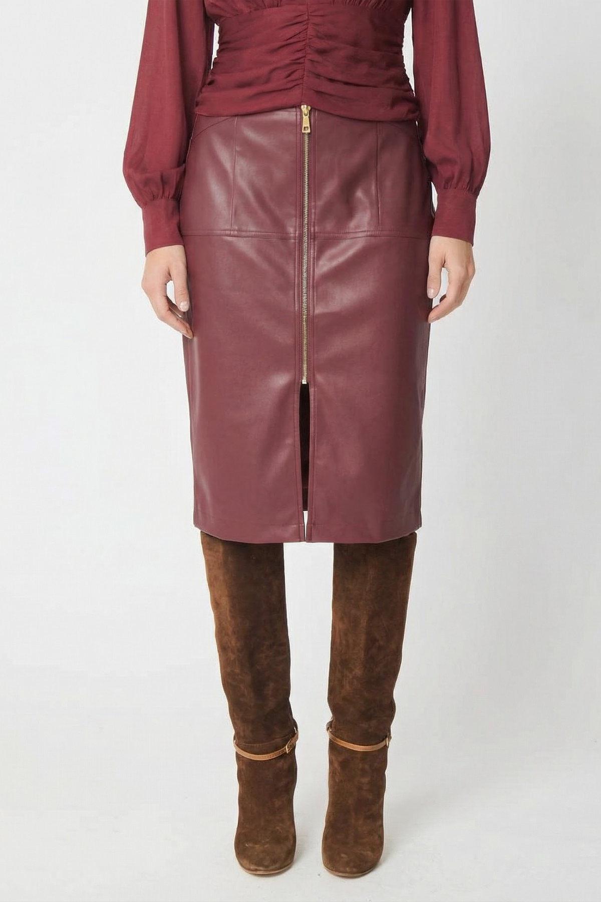 A model is shown from the chest down wearing a knee-length burgundy faux-leather skirt with a front zipper, paired with a matching burgundy top and brown suede over-the-knee boots, against a plain white studio background.