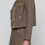 A model wearing a cropped tweed blazer and matching shorts in taupe-brown with prominent gold buttons, shown from the side against a light gray studio background.