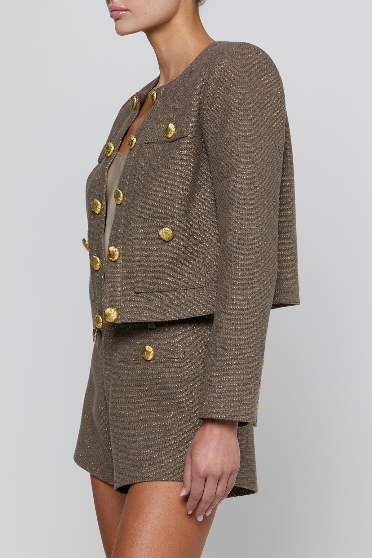 A model wearing a cropped tweed blazer and matching shorts in taupe-brown with prominent gold buttons, shown from the side against a light gray studio background.