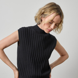 A woman wearing a black ribbed sleeveless mock-neck sweater vest, shown from the waist up against a plain light gray studio background.