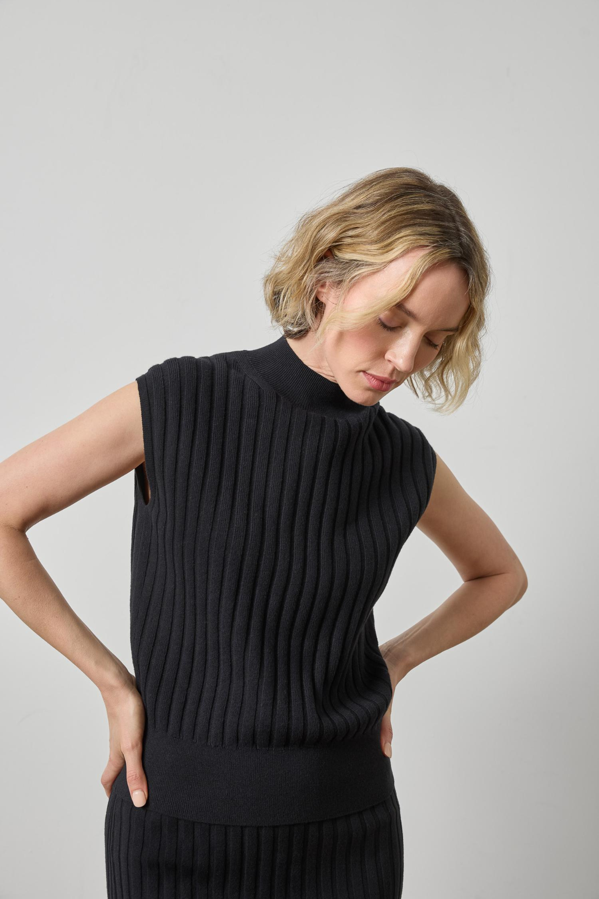 A woman wearing a black ribbed sleeveless mock-neck sweater vest, shown from the waist up against a plain light gray studio background.