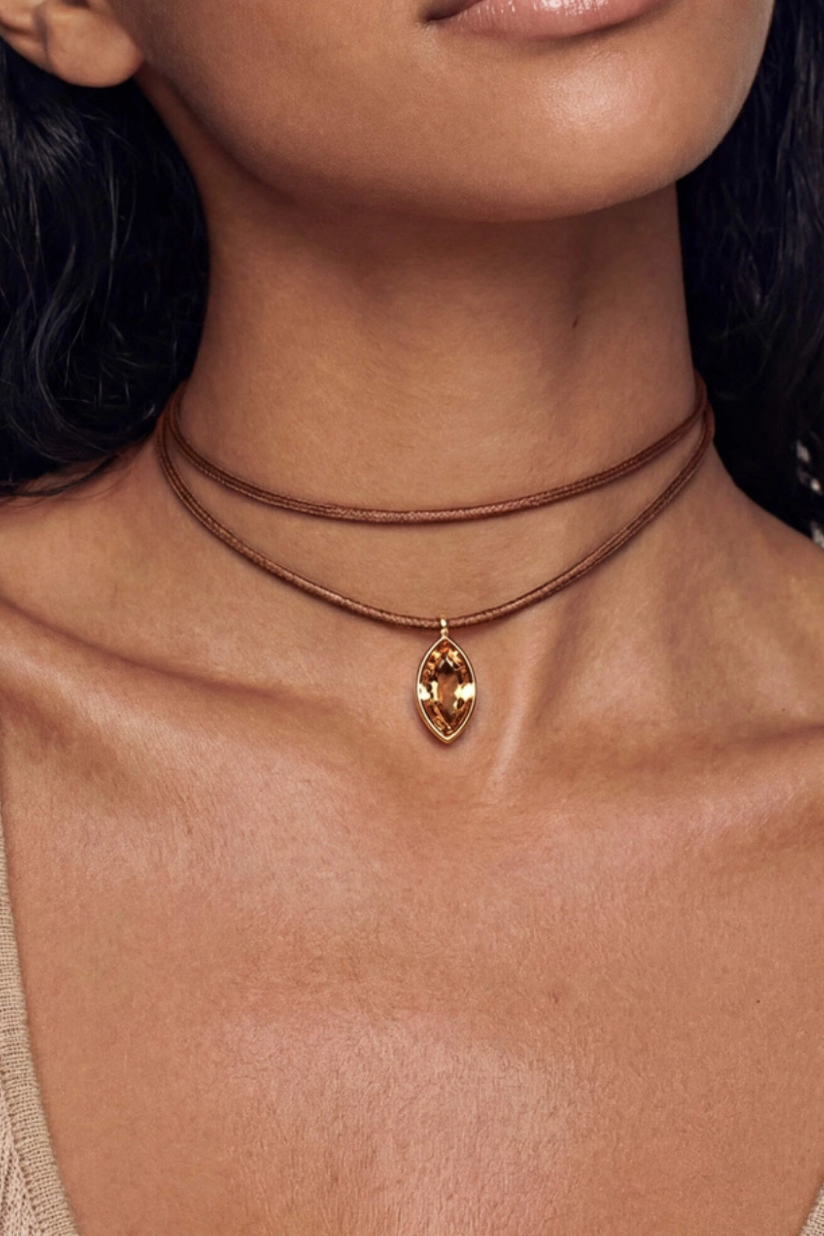 A close-up of a model wearing a double-strand brown cord choker necklace with a gold-toned marquise-shaped pendant containing a faceted amber gemstone.