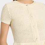 Close-up of a model wearing a cream-colored short-sleeve knit top with a textured openwork pattern, decorative buttons, and a fringed hem.
