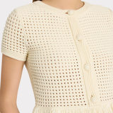 Close-up of a model wearing a cream-colored short-sleeve knit top with a textured openwork pattern, decorative buttons, and a fringed hem.
