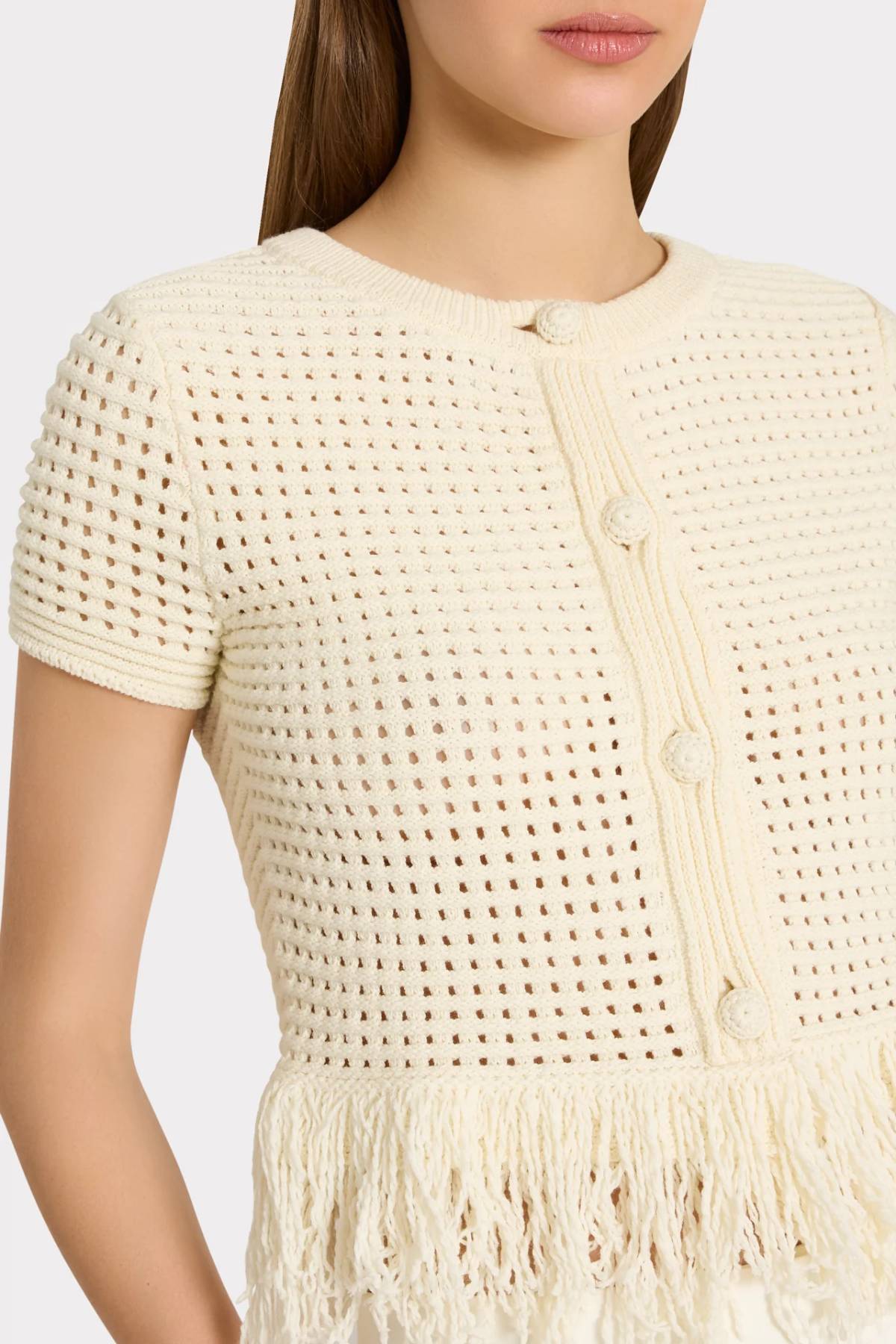 Close-up of a model wearing a cream-colored short-sleeve knit top with a textured openwork pattern, decorative buttons, and a fringed hem.