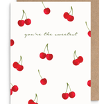 A white greeting card with a pattern of red cherries and the phrase 'you're the sweetest' in green script, shown with a brown kraft envelope behind it on a white background.