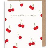 A white greeting card with a pattern of red cherries and the phrase 'you're the sweetest' in green script, shown with a brown kraft envelope behind it on a white background.