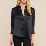 A blonde model wearing a black satin button-down shirt with three-quarter sleeves and black pants, standing against a solid beige background.