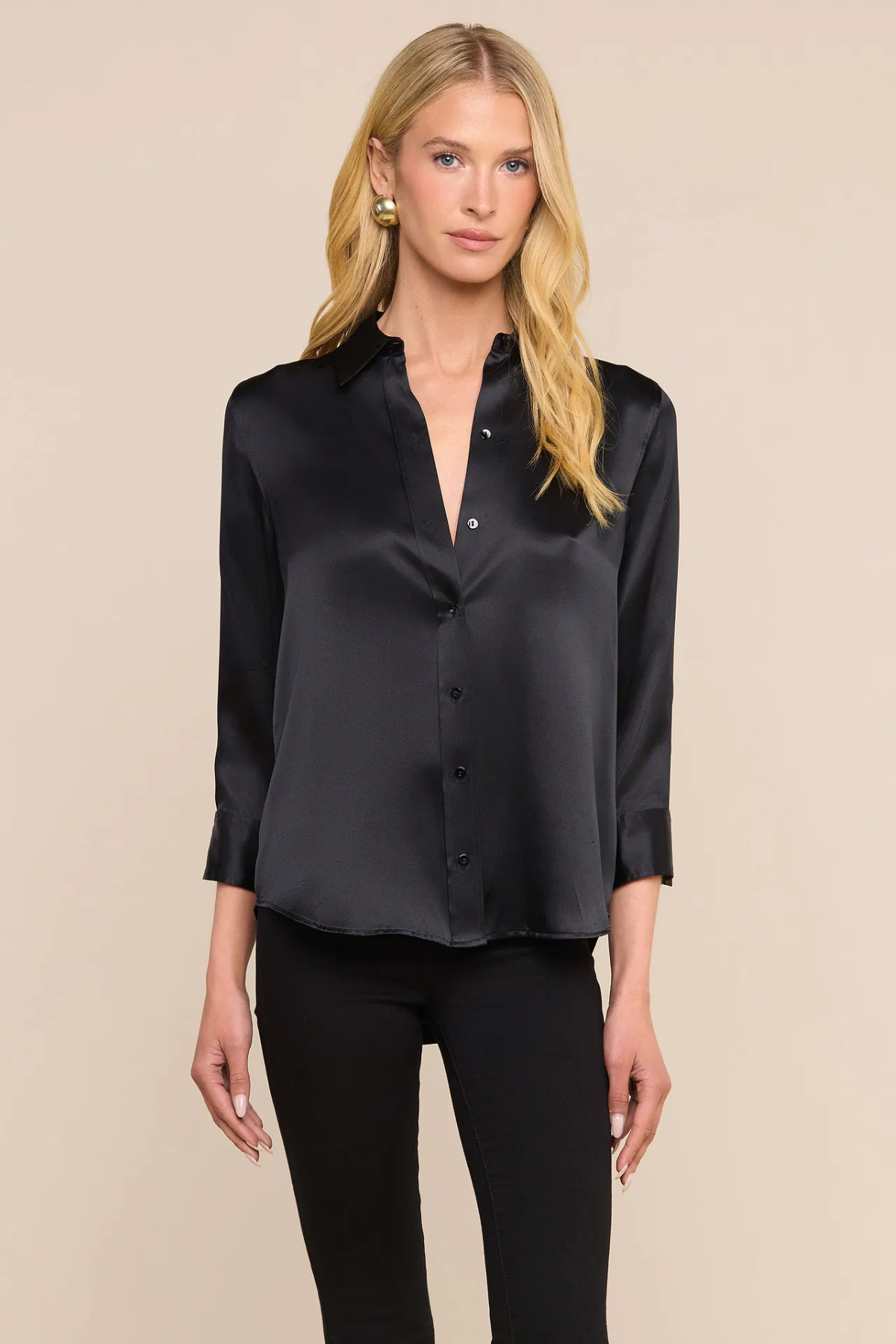 A blonde model wearing a black satin button-down shirt with three-quarter sleeves and black pants, standing against a solid beige background.