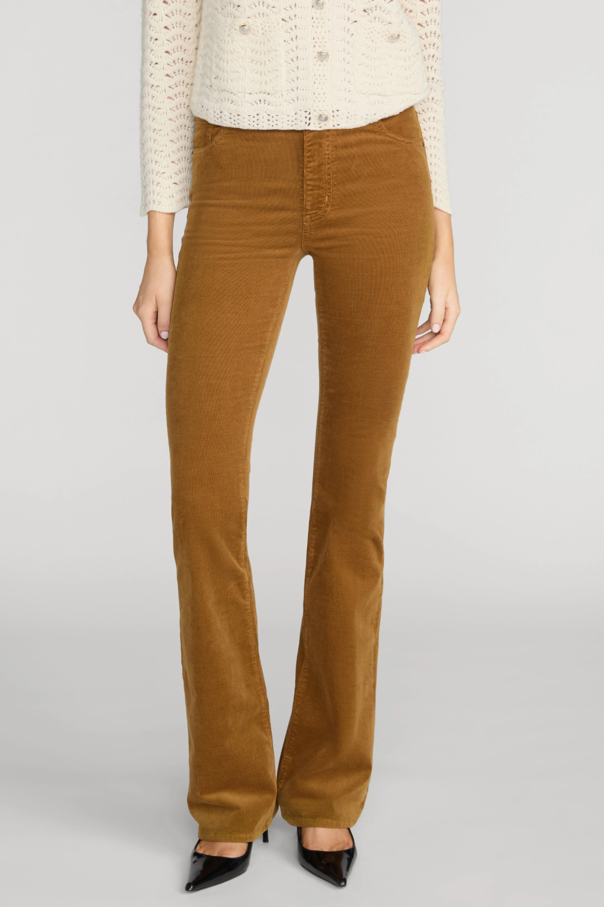 A model wearing a pair of brown corduroy flare pants with a cream knit top and black pointed-toe heels, shown from the chest down against a light gray studio background.
