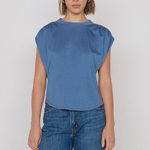 A woman modeling a blue short-sleeve top with gathered shoulder details and blue jeans against a plain light gray studio background.