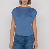 A woman modeling a blue short-sleeve top with gathered shoulder details and blue jeans against a plain light gray studio background.