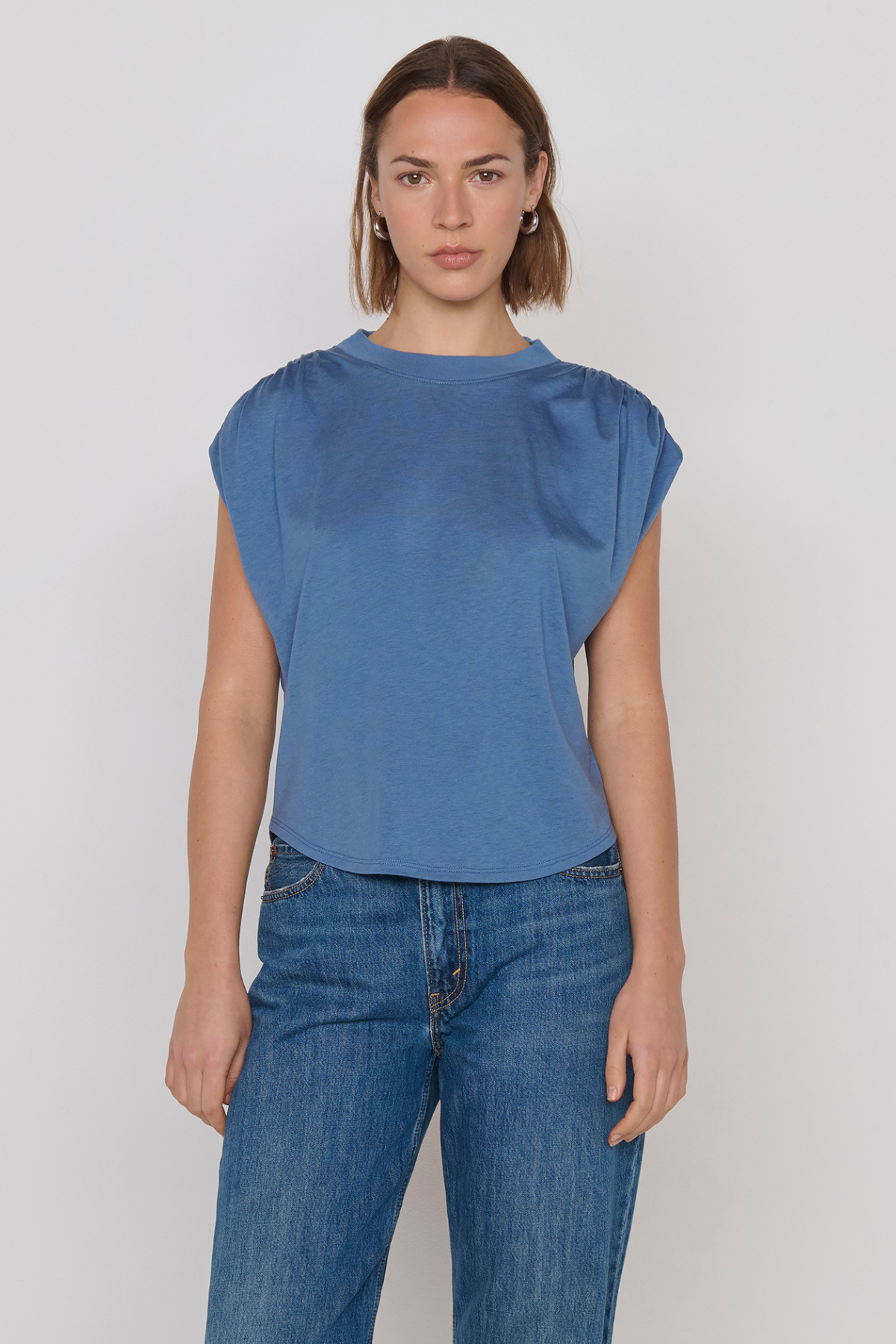 A woman modeling a blue short-sleeve top with gathered shoulder details and blue jeans against a plain light gray studio background.