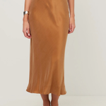 A woman wearing a high-waisted, midi-length satin slip skirt in camel brown, paired with a navy top and black pointed flats, photographed against a neutral light gray studio background.