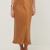 A woman wearing a high-waisted, midi-length satin slip skirt in camel brown, paired with a navy top and black pointed flats, photographed against a neutral light gray studio background.