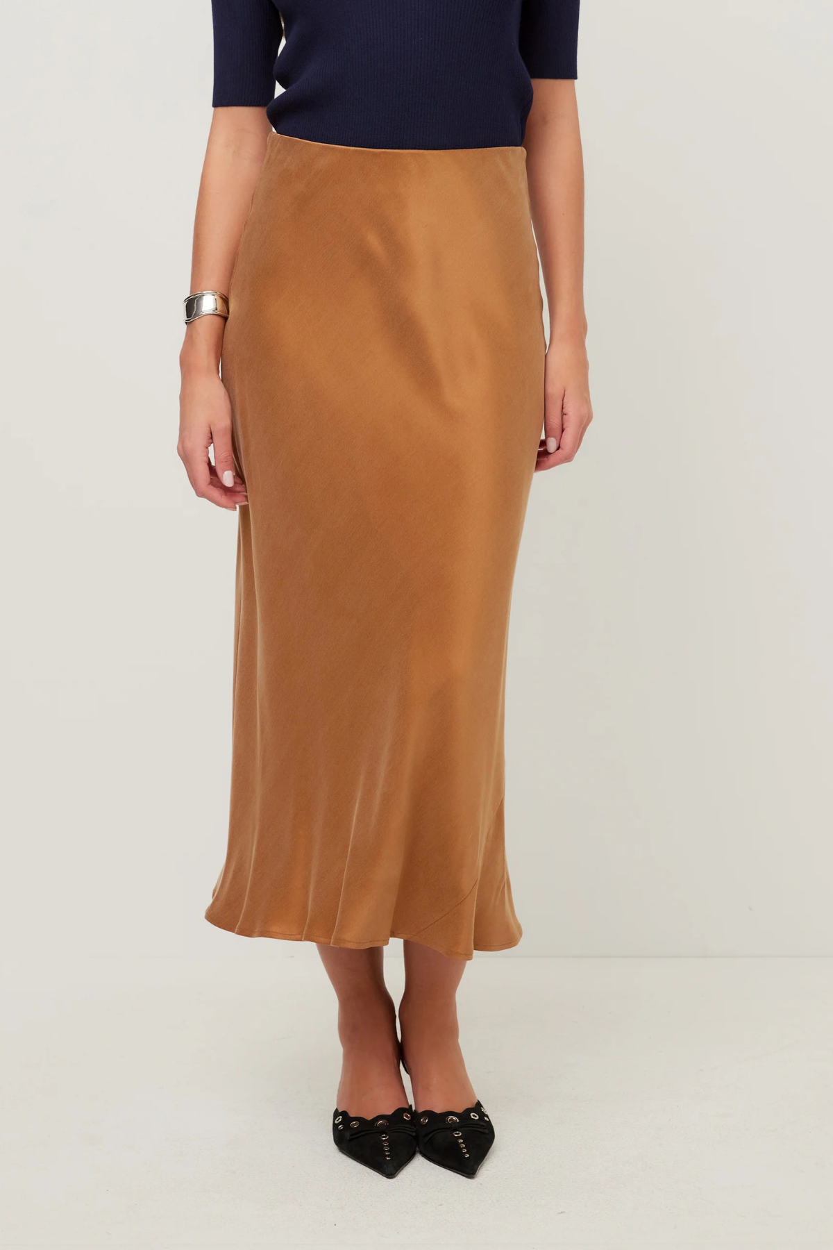 A woman wearing a high-waisted, midi-length satin slip skirt in camel brown, paired with a navy top and black pointed flats, photographed against a neutral light gray studio background.