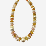 A beaded necklace featuring a mix of polished gold, amber, pink, and cream-colored stones, with a prominent textured gold bead at the center, displayed on a light gray background.