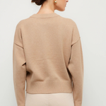 A woman shown from the back wearing a light beige crewneck knit sweater and light beige pants against a plain light gray studio background.