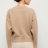 A woman shown from the back wearing a light beige crewneck knit sweater and light beige pants against a plain light gray studio background.