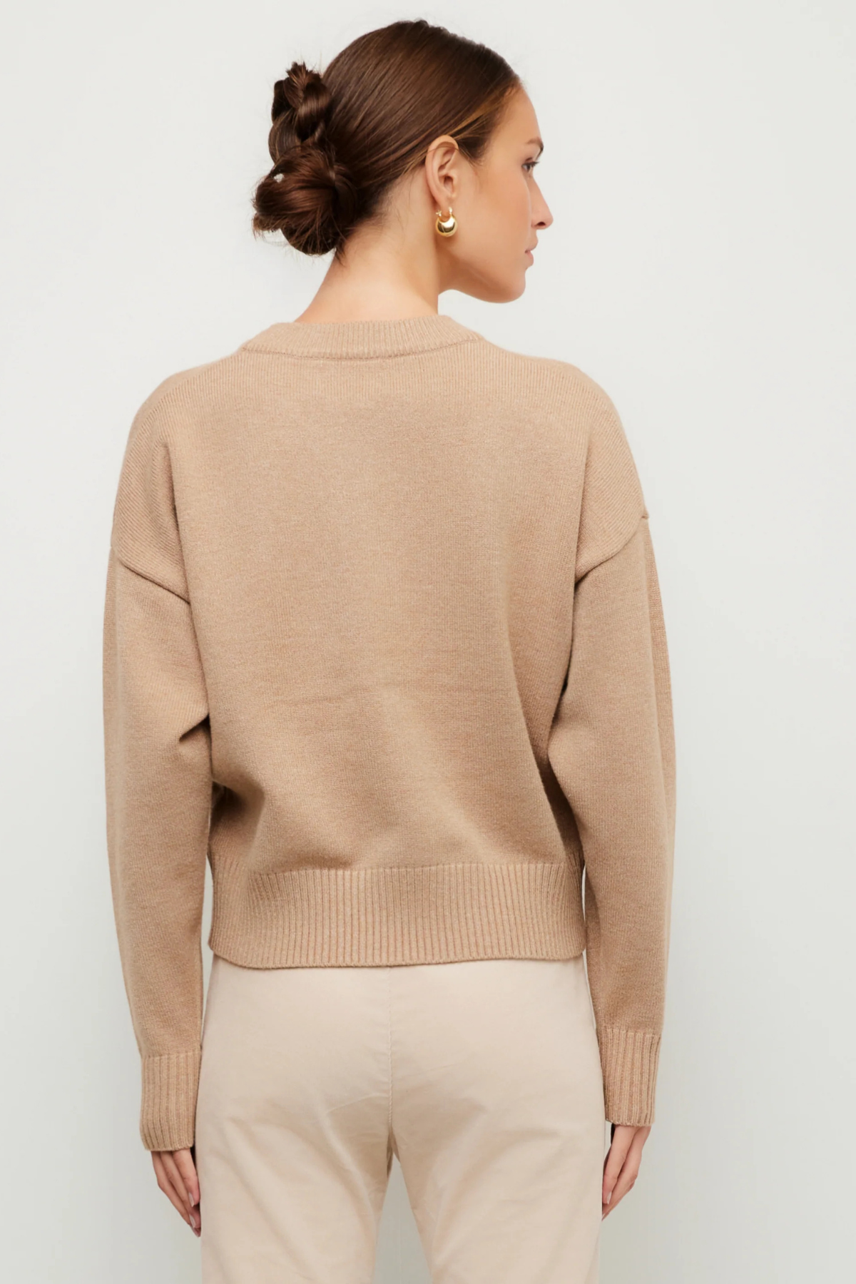 A woman shown from the back wearing a light beige crewneck knit sweater and light beige pants against a plain light gray studio background.