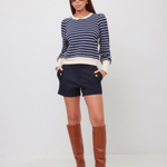 A woman modeling a navy and white striped long-sleeve knit top, navy shorts, and brown knee-high leather boots against a plain light gray studio background.