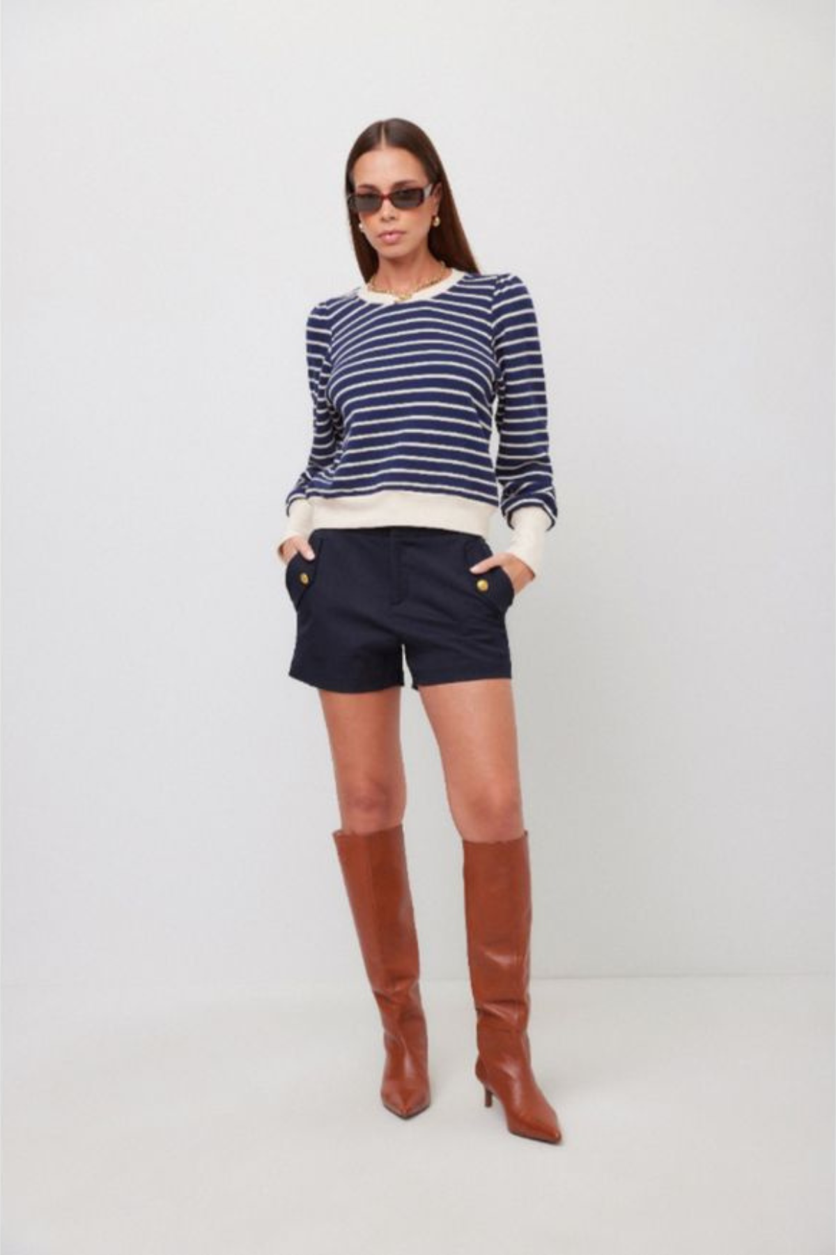 A woman modeling a navy and white striped long-sleeve knit top, navy shorts, and brown knee-high leather boots against a plain light gray studio background.