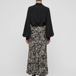 A model shown from the back wearing a black long-sleeve blouse and a black midi skirt with a beige floral pattern, paired with black pointed-toe heels, against a light gray studio background.