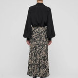 A model shown from the back wearing a black long-sleeve blouse and a black midi skirt with a beige floral pattern, paired with black pointed-toe heels, against a light gray studio background.