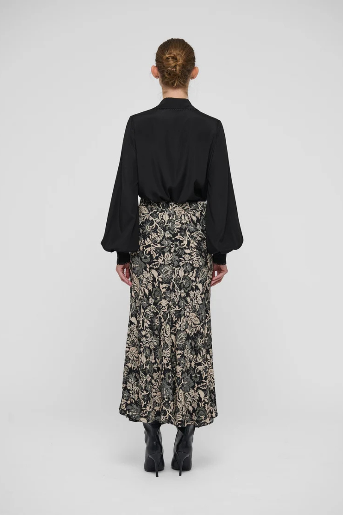 A model shown from the back wearing a black long-sleeve blouse and a black midi skirt with a beige floral pattern, paired with black pointed-toe heels, against a light gray studio background.