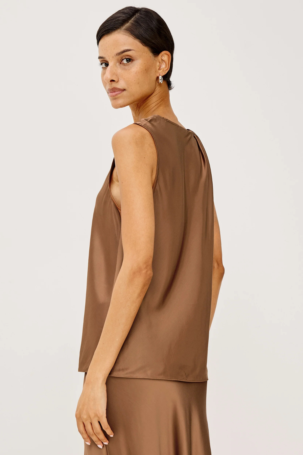A woman modeling a sleeveless taupe-brown satin top, shown from a side-back angle against a clean light gray studio background.