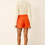 back of woman in white knit top and orange shorts on white background 