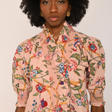 A model with a large black afro wearing a pink short-sleeve button-up shirt with a colorful floral and bird print, paired with dark brown pants, against a plain light background.