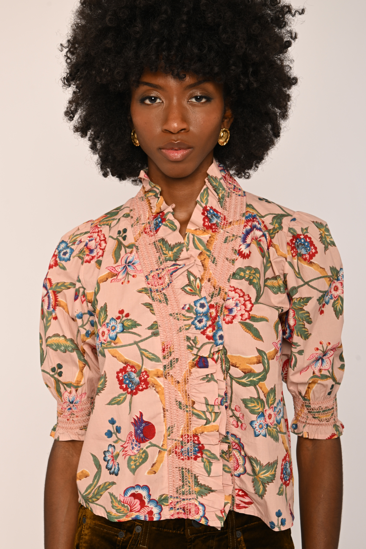 A model with a large black afro wearing a pink short-sleeve button-up shirt with a colorful floral and bird print, paired with dark brown pants, against a plain light background.