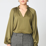 A woman modeling a long-sleeve olive green blouse with a gathered ruffled neckline, paired with gray pleated trousers, against a light gray studio background.