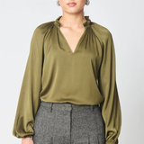 A woman modeling a long-sleeve olive green blouse with a gathered ruffled neckline, paired with gray pleated trousers, against a light gray studio background.