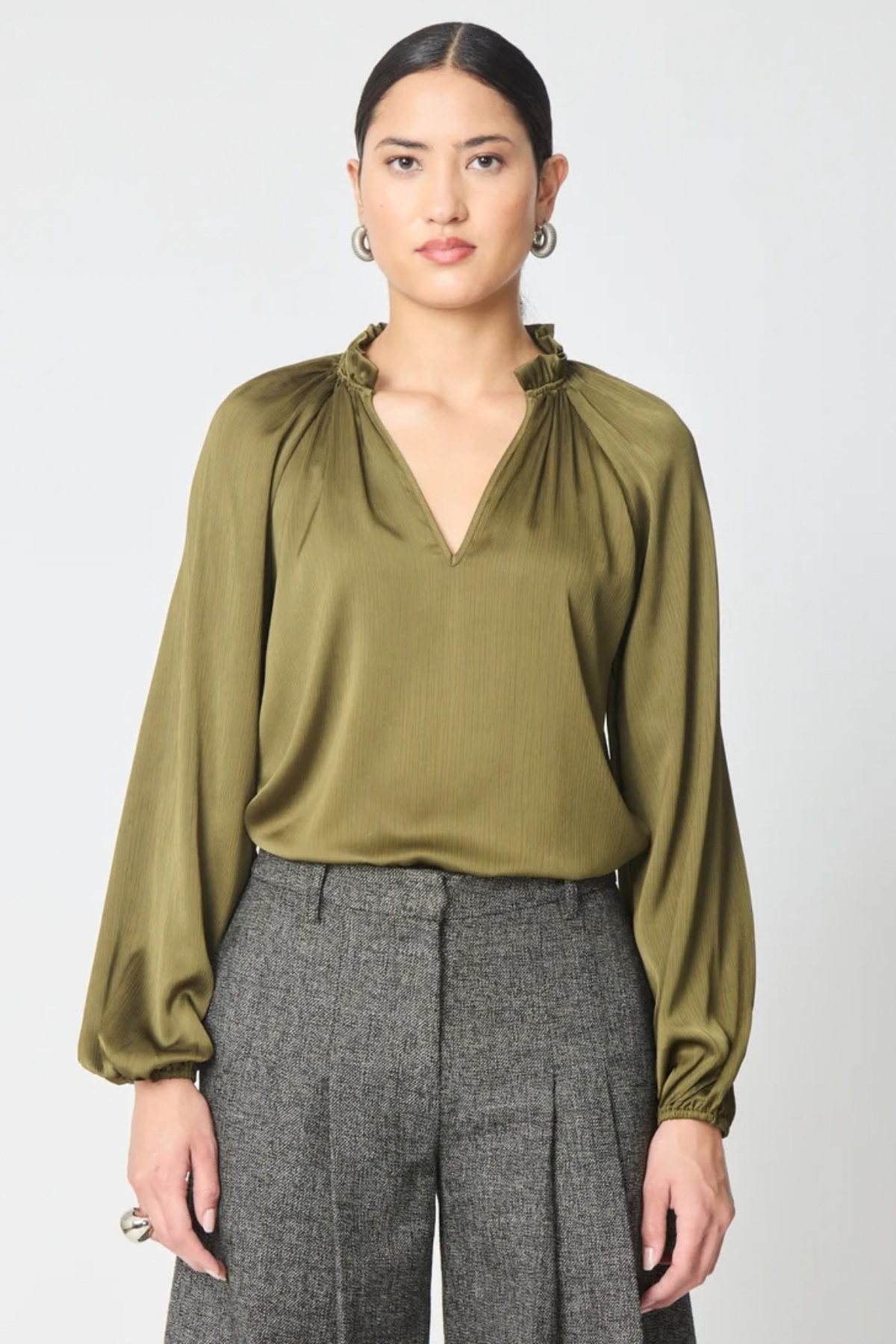A woman modeling a long-sleeve olive green blouse with a gathered ruffled neckline, paired with gray pleated trousers, against a light gray studio background.