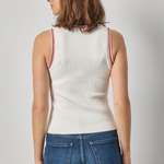 Woman wearing a white ribbed tank top with red trim and blue jeans on a gray background