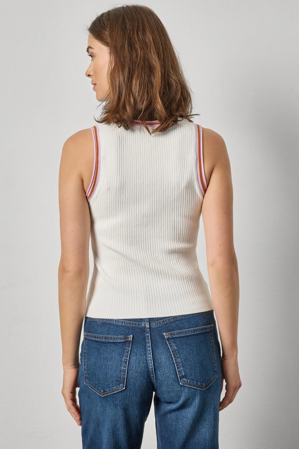 Woman wearing a white ribbed tank top with red trim and blue jeans on a gray background