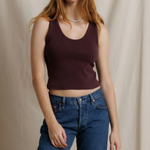 A woman modeling a dark burgundy ribbed tank top paired with blue jeans, standing against a draped beige fabric backdrop.